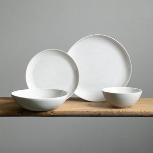 Four different types of plate on a wooden table. You can see a small and large plate placed on top of each other, in front of which is a pasta plate and a muesli bowl in white glaze with fine golden spots.