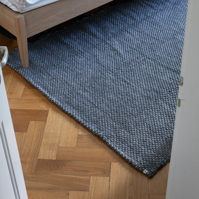 Gray large carpet hand-woven pattern lies in front of a bed 