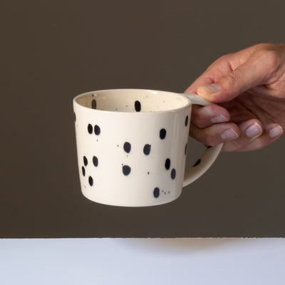 The cream-colored tea cup with black splashes of color is held by a hand on the large handle.