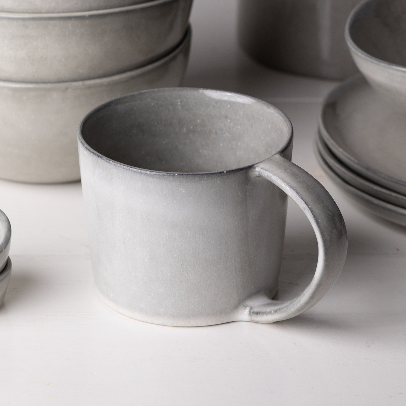 In the foreground is a large teacup with a handle with a light gray reactive glaze made of stoneware in the background are large plates and muesli bowls