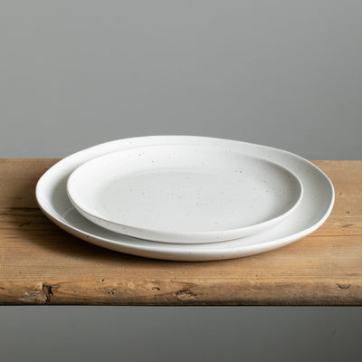 A small plate lies on a large plate, both have an organic shape and are white with golden brown spots