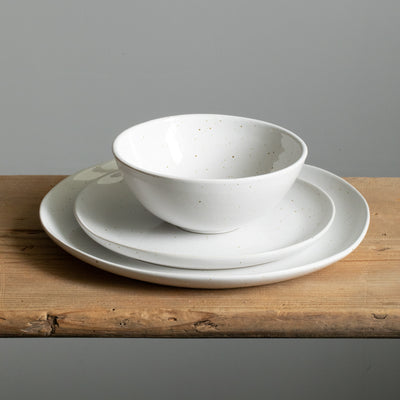 A cereal bowl stands on a small breakfast plate underneath which is a large organic main course plate in glossy white with fine golden-brown sprinkles