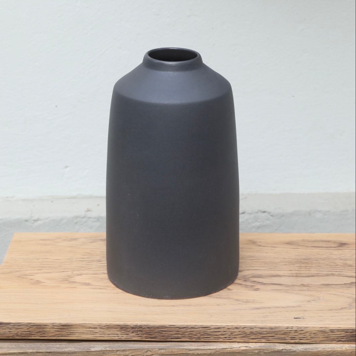 The matt black vase with a small opening at the top and a wide lower section stands on a wooden board.
