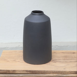 The matt black vase with a small opening at the top and a wide lower section stands on a wooden board.